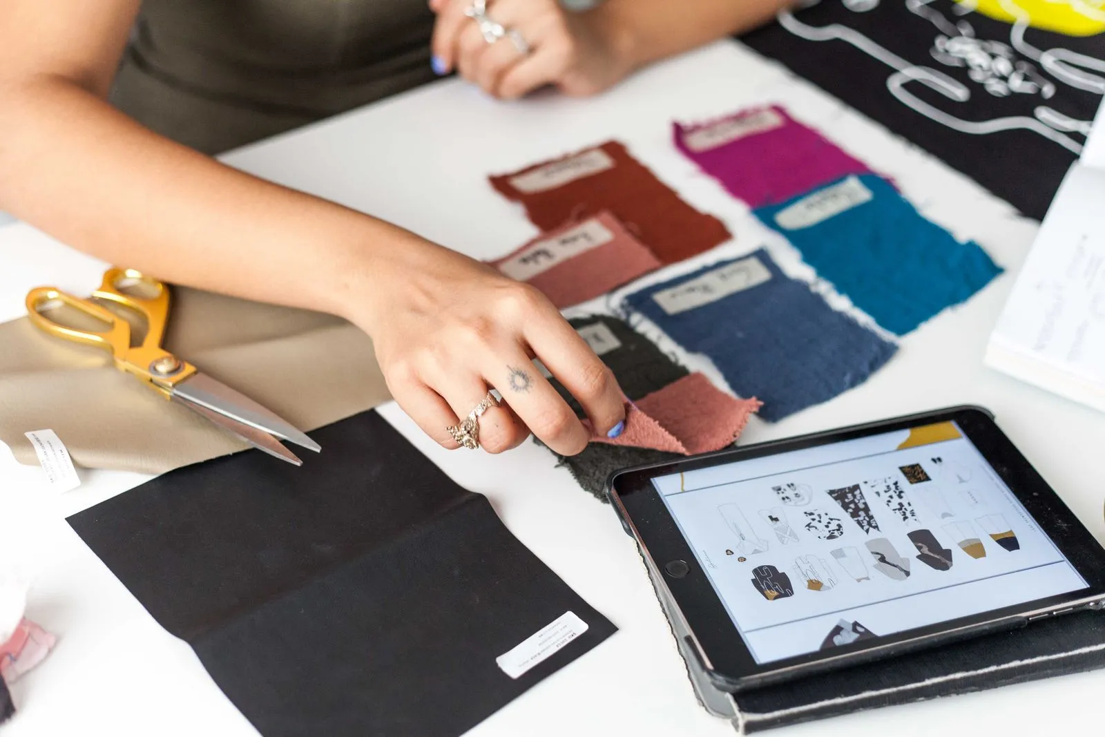 Fashion designer examines fabric swatches and digital designs on tablet indoors.